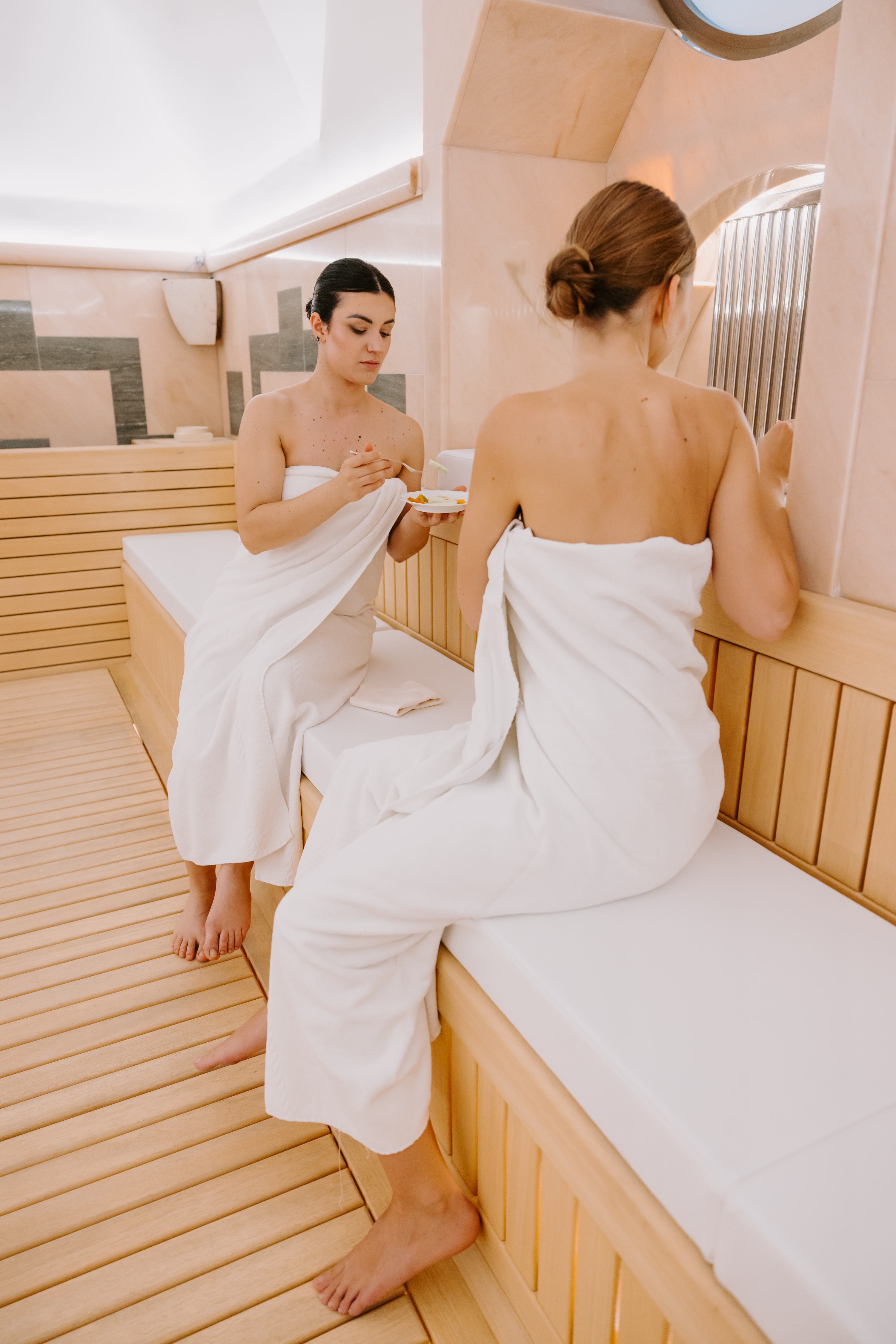 Two women in sauna with white towels drinking herbal tea